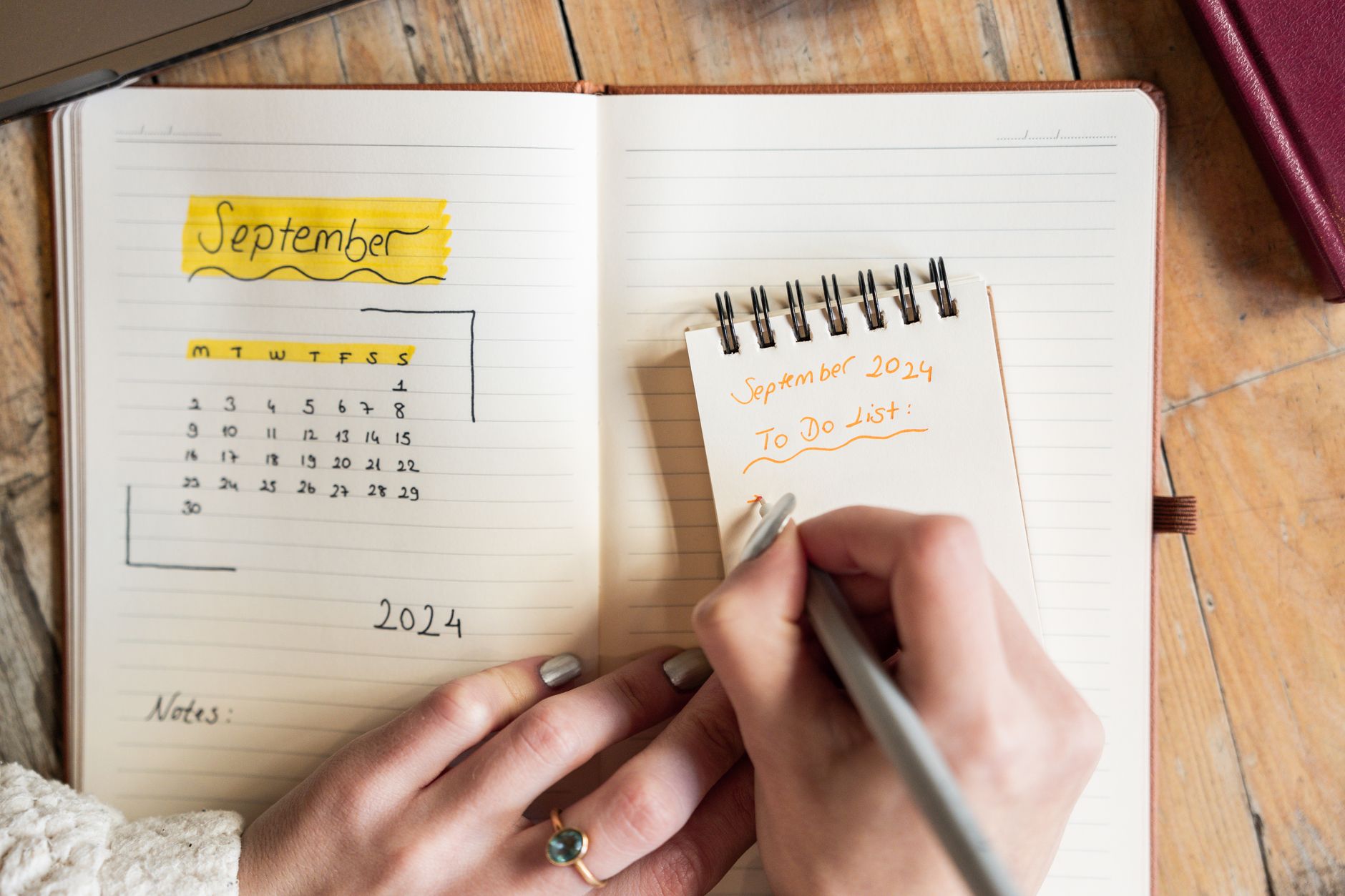 Hands writing a to-do list on a small spiral notepad resting on an open planner.