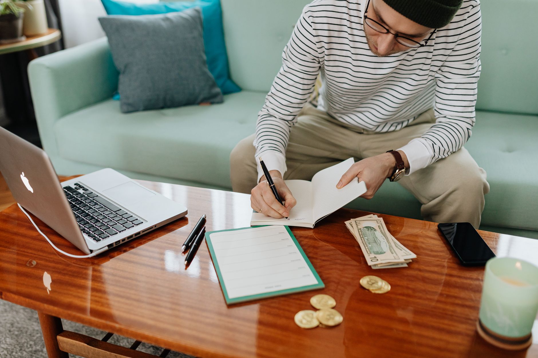 A man in a striped shirt sitting on a sofa, writing in a notebook at a coffee table with a laptop beside him.